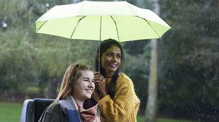 Woman holding a green umbrella over another woman in a wheelchair, both smiling out to the left